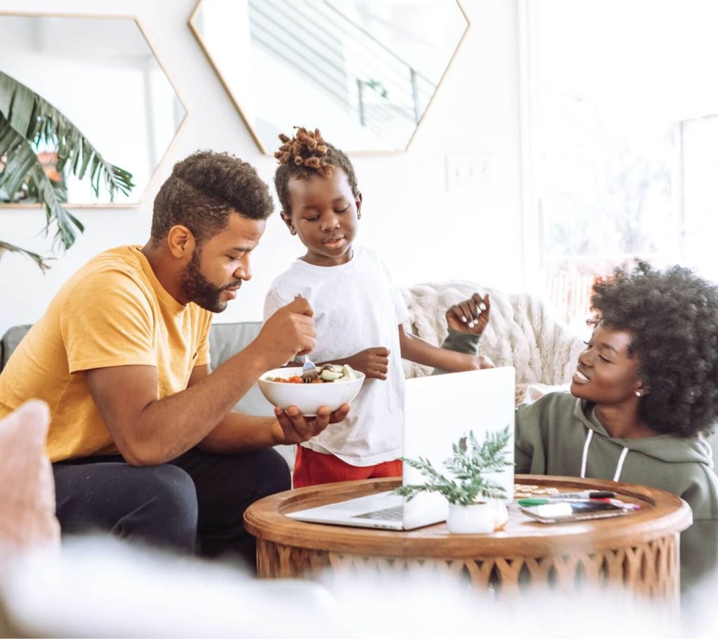 A father holding a bowl of food with a boy standing next him while the mother sits on the floor behind a coffee table